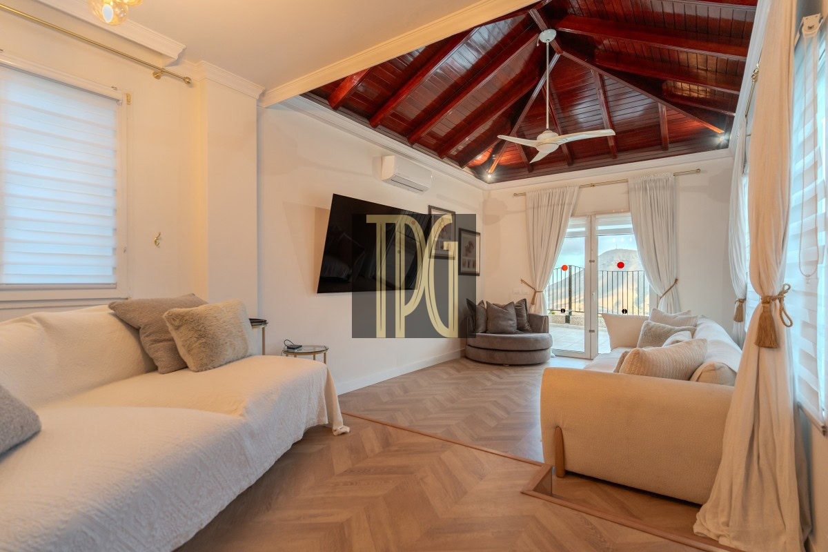 Bright living room with a white sofa set, beige armchairs, and a wooden vaulted ceiling with a ceiling fan and large windows leads to a balcony.