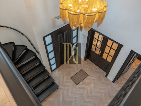 Two-story foyer with a black metal staircase on the left, dark double doors with glass panes, and a warm chandelier overhead above a herringbone floor.