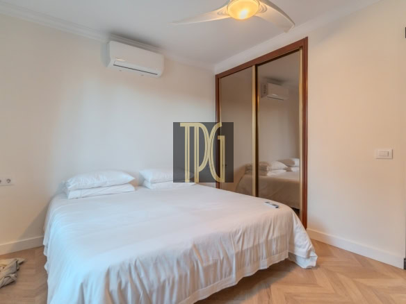 Bedroom with white bedding, two pillows, a ceiling fan, and a wall-mounted air conditioner; a large wooden-framed mirrored wardrobe reflects the room