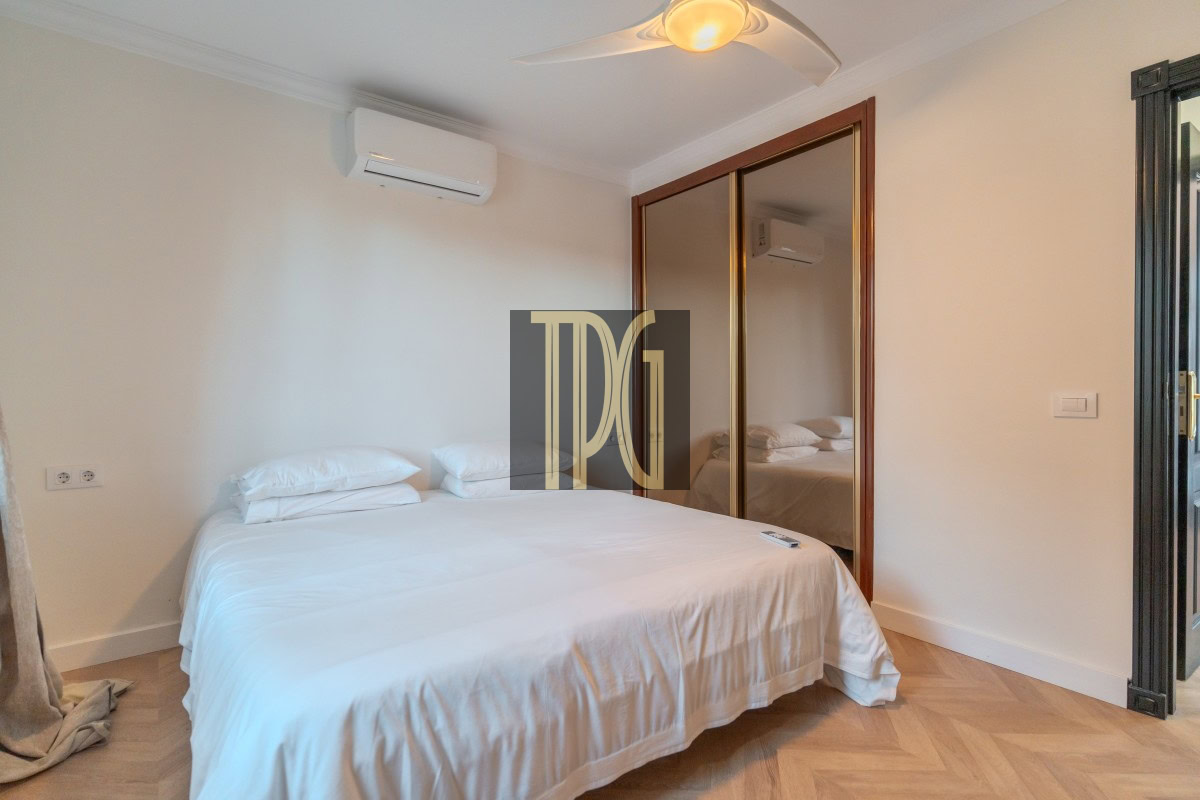 Bedroom with white bedding, two pillows, a ceiling fan, and a wall-mounted air conditioner; a large wooden-framed mirrored wardrobe reflects the room