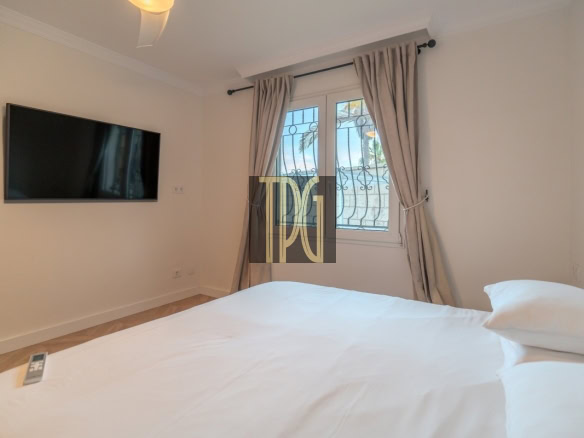 Bedroom with a large white bed, a flat-screen TV on the wall, and a window with beige curtains.