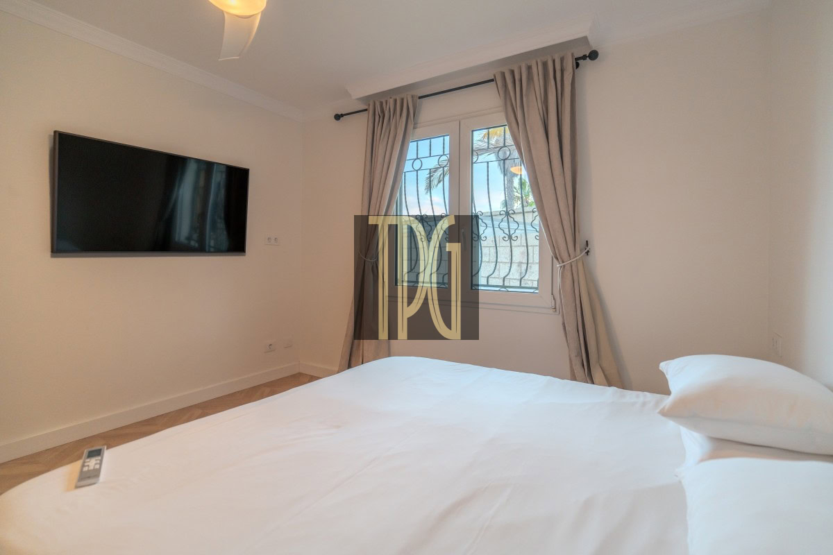 Bedroom with a large white bed, a flat-screen TV on the wall, and a window with beige curtains.