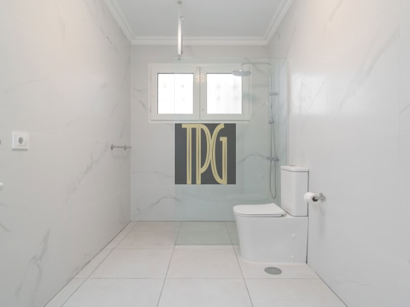 Bright, minimalist white bathroom with large marble-like tiles, glass shower enclosure, wall-mounted toilet, and a window at the far wall, bathed in natural light.