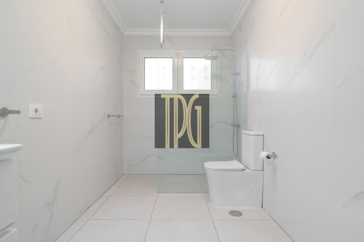 Bright, minimalist white bathroom with large marble-like tiles, glass shower enclosure, wall-mounted toilet, and a window at the far wall, bathed in natural light.