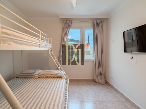 Small white bunk bed room with a ladder, striped gray‑and‑white sheets, a window with beige curtains, and a wall‑mounted TV.