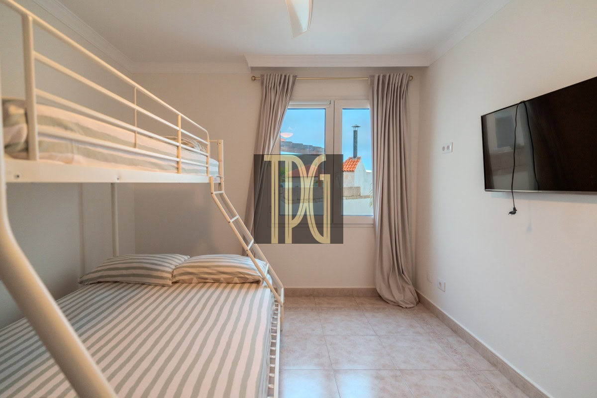 Small white bunk bed room with a ladder, striped gray‑and‑white sheets, a window with beige curtains, and a wall‑mounted TV.