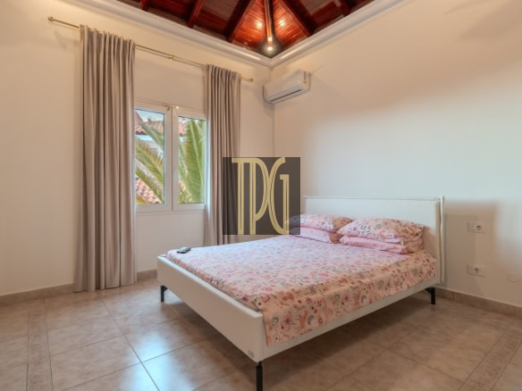 White upholstered bed with pink floral bedding in a bright bedroom, beige tiled floor, and a large window with light curtains showing palm trees outside, plus a wooden vaulted ceiling and wall-mounted AC above the bed.