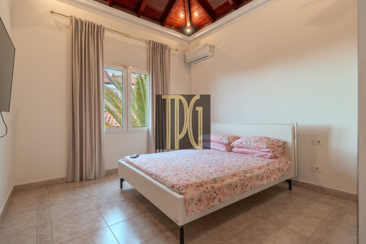 White upholstered bed with pink floral bedding in a bright bedroom, beige tiled floor, and a large window with light curtains showing palm trees outside, plus a wooden vaulted ceiling and wall-mounted AC above the bed.