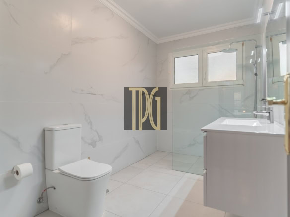 Modern white bathroom with a glass shower enclosure, wall-mounted vanity, and a white toilet set against marble-patterned walls and tiled floor to the left and a frosted window above the shower on the right.