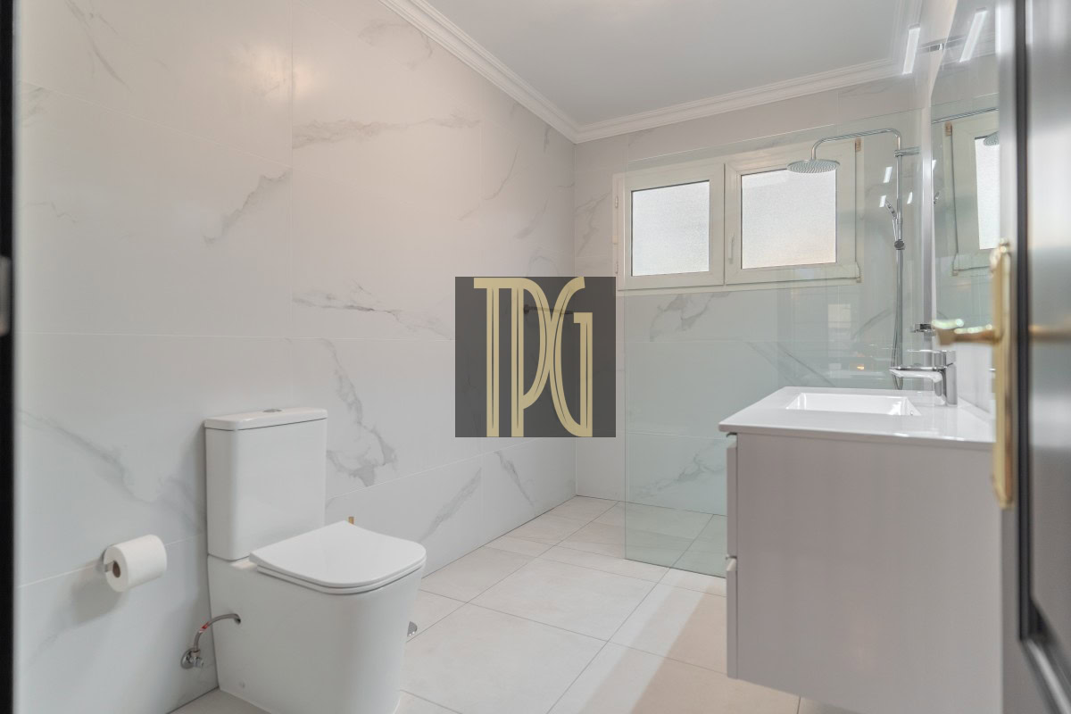 Modern white bathroom with a glass shower enclosure, wall-mounted vanity, and a white toilet set against marble-patterned walls and tiled floor to the left and a frosted window above the shower on the right.