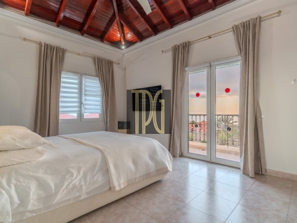Bright bedroom with white bedding, beige curtains, tiled floor, a wood vaulted ceiling, and glass doors to a balcony with a sunset view.