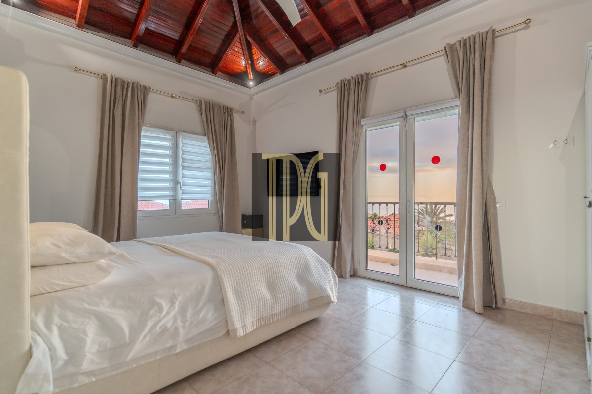 Bright bedroom with white bedding, beige curtains, tiled floor, a wood vaulted ceiling, and glass doors to a balcony with a sunset view.