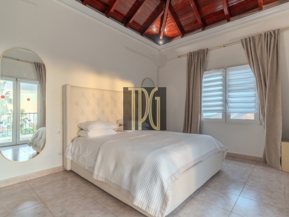 Bright bedroom with a white tufted headboard bed, white linens, and light tiled floor; a large oval wall mirror on the left and beige curtains by a window to the right.
