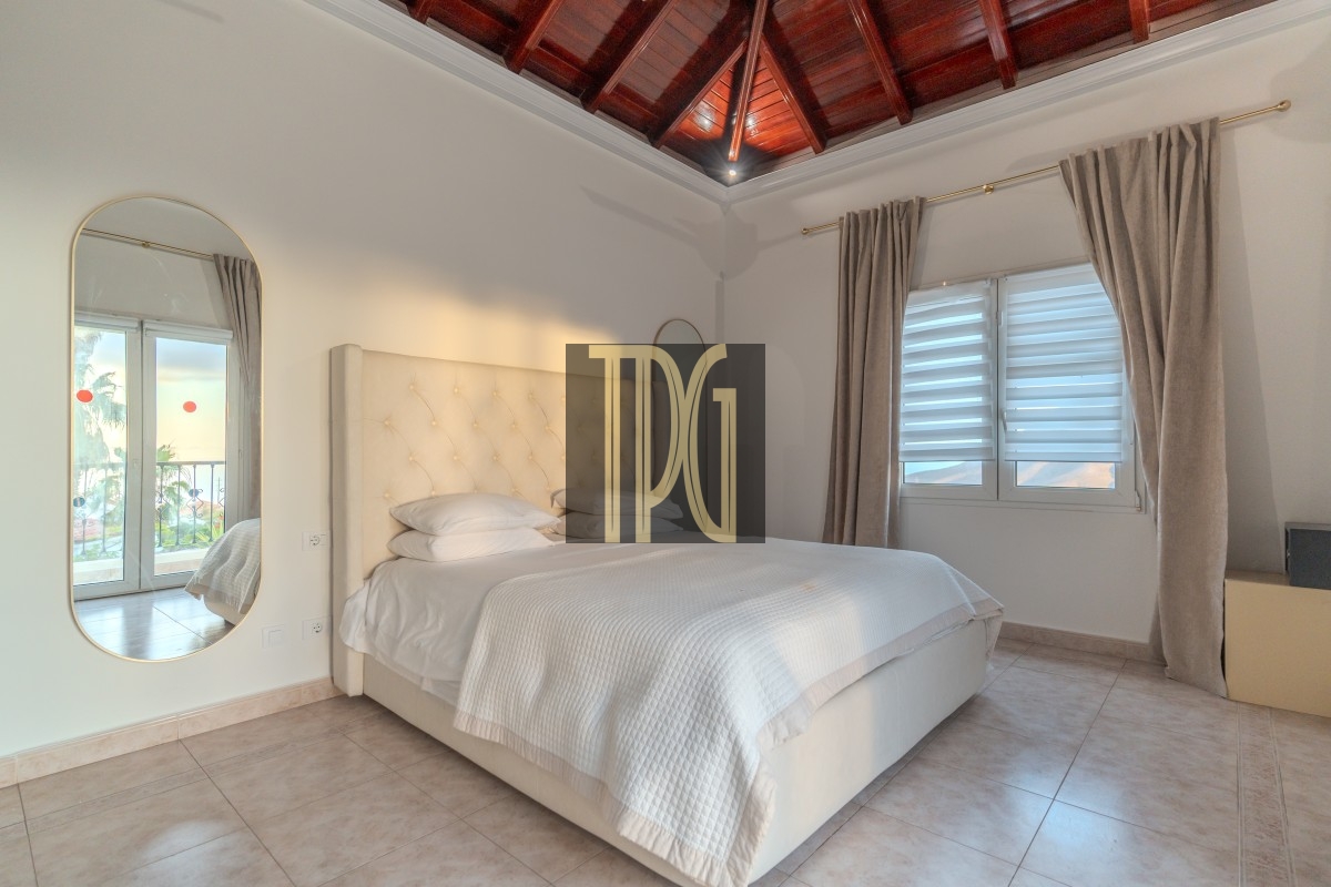 Bright bedroom with a white tufted headboard bed, white linens, and light tiled floor; a large oval wall mirror on the left and beige curtains by a window to the right.