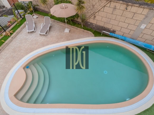 Backyard pool area with curved steps, beige coping, and two white lounge chairs under a tan umbrella on a tiled patio.