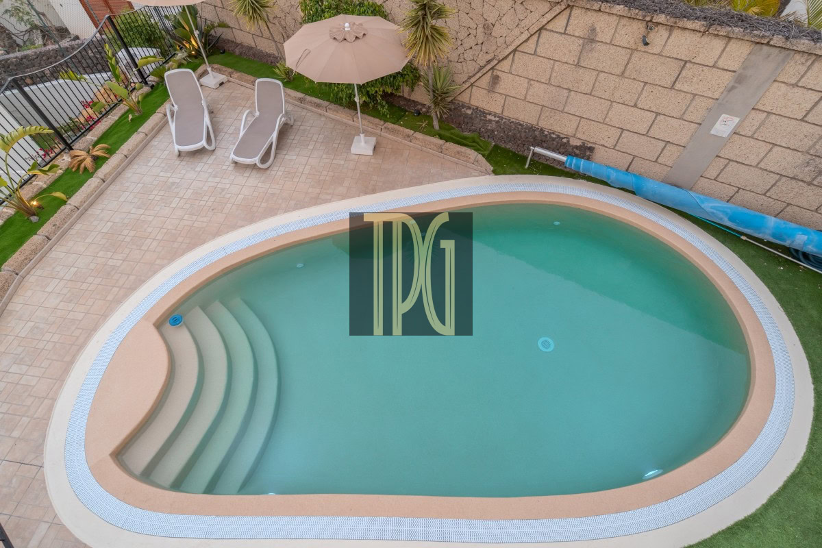 Backyard pool area with curved steps, beige coping, and two white lounge chairs under a tan umbrella on a tiled patio.