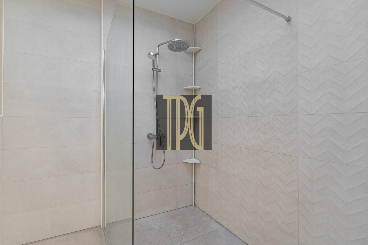 Modern walk-in shower with a glass door, a ceiling rain showerhead and handheld wand, Beige tiled walls with corner shelves.
