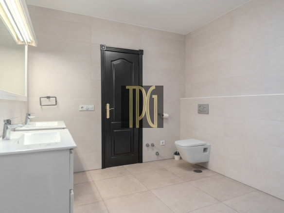 Modern beige-tiled bathroom featuring a black paneled door, white double vanity with sinks on the left, and a wall-mounted toilet on the right across from the door. A small plant sits near the toilet wall.