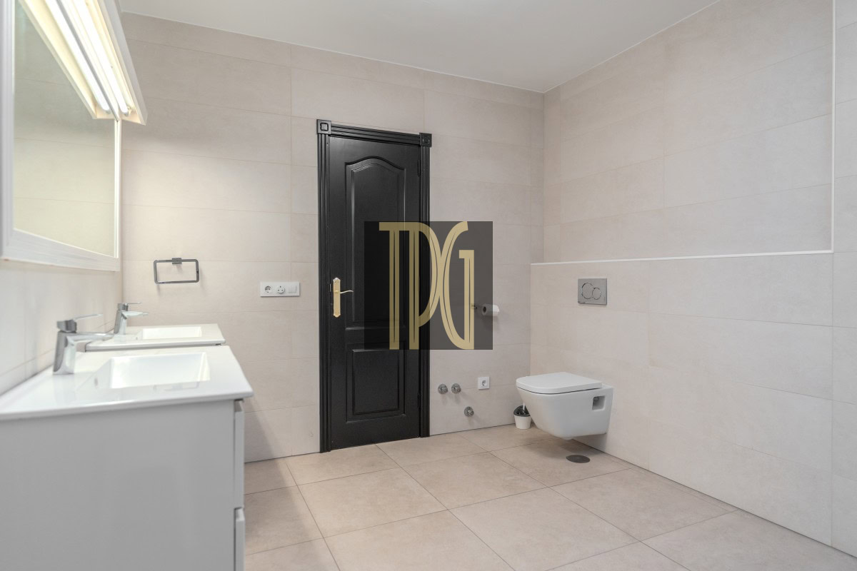 Modern beige-tiled bathroom featuring a black paneled door, white double vanity with sinks on the left, and a wall-mounted toilet on the right across from the door. A small plant sits near the toilet wall.