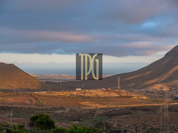 Desert hillside landscape at dusk with rolling brown hills, sparse vegetation, and a distant coastline on the horizon under a cloudy sky.