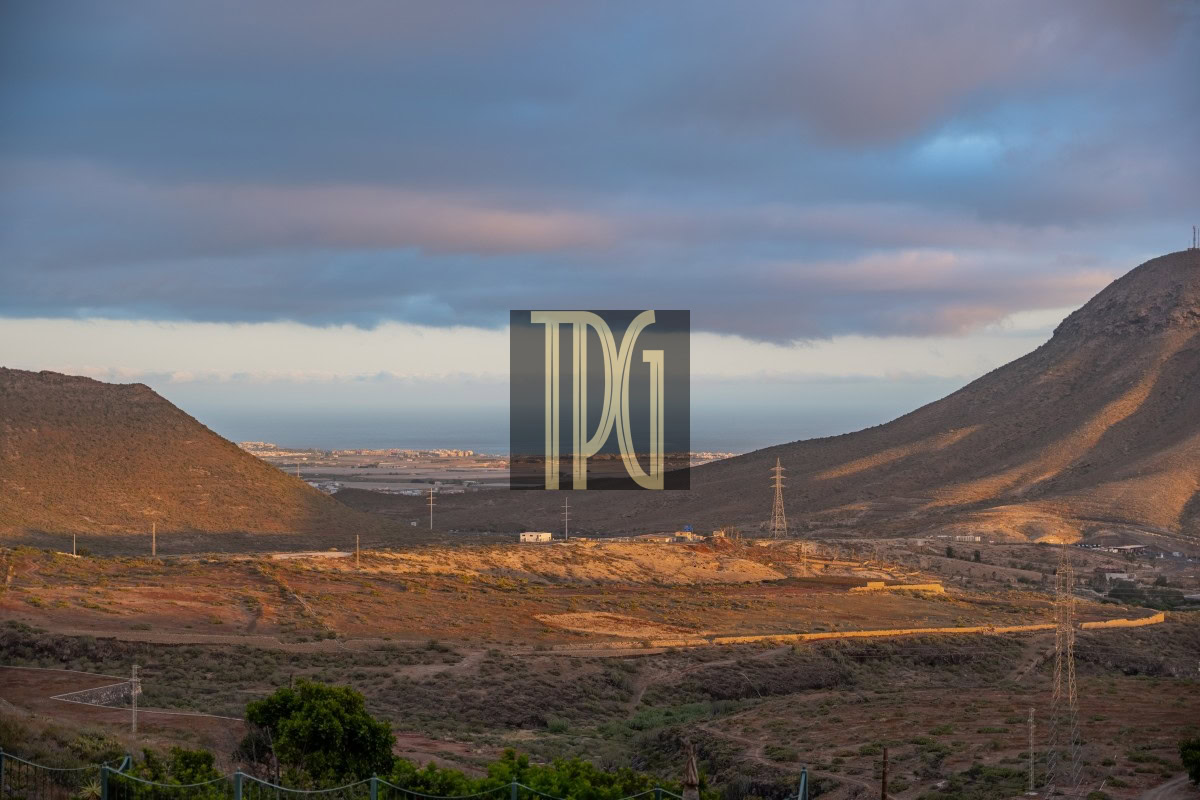 Desert hillside landscape at dusk with rolling brown hills, sparse vegetation, and a distant coastline on the horizon under a cloudy sky.