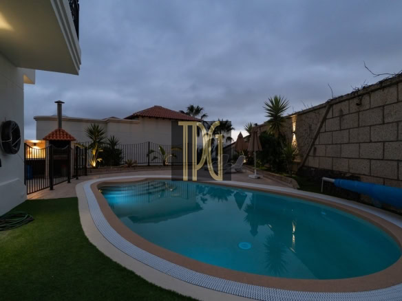 Oval backyard swimming pool at dusk, fenced patio with lounge chairs and umbrellas, palm plants and a stone wall on the right.