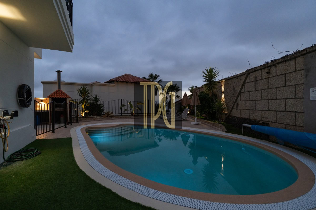 Oval backyard swimming pool at dusk, fenced patio with lounge chairs and umbrellas, palm plants and a stone wall on the right.