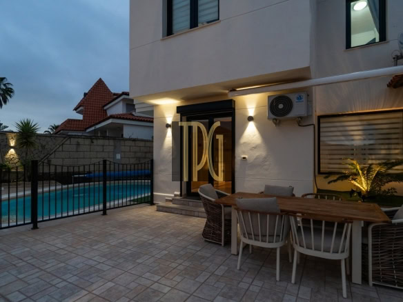 Outdoor patio at dusk with a rectangular pool, black fence, and a wooden dining table surrounded by cushioned chairs beside a modern house entrance.