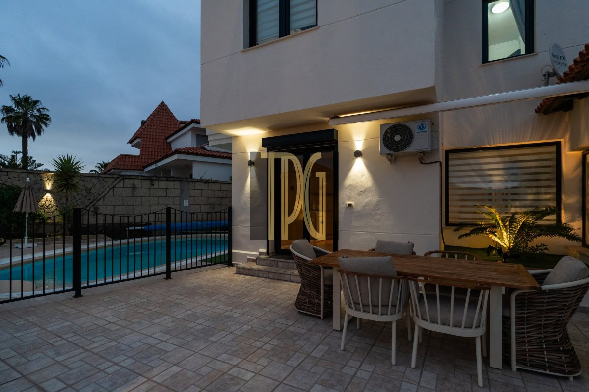 Outdoor patio at dusk with a rectangular pool, black fence, and a wooden dining table surrounded by cushioned chairs beside a modern house entrance.