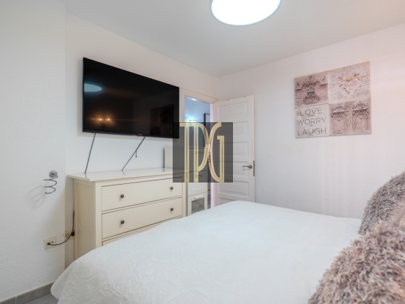 Bedroom with a wall-mounted TV above a white dresser, open door, and a quilted white bed with fluffy pillows in soft lighting.