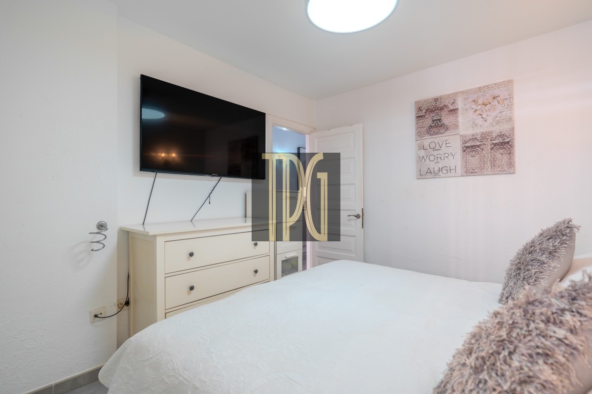 Bedroom with a wall-mounted TV above a white dresser, open door, and a quilted white bed with fluffy pillows in soft lighting.