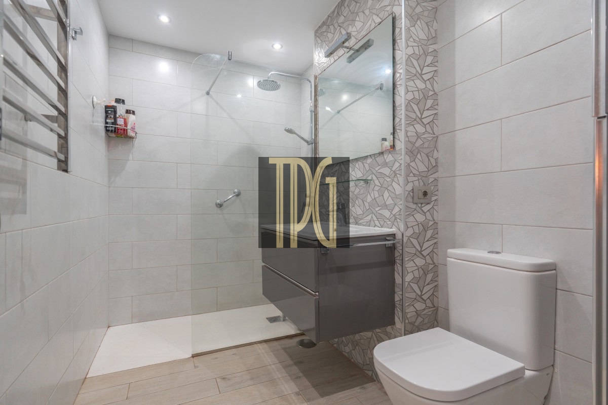 Modern bathroom with glass-enclosed shower, rainfall head, and a floating gray vanity with mirror and geometric tile accent wall on the right side.