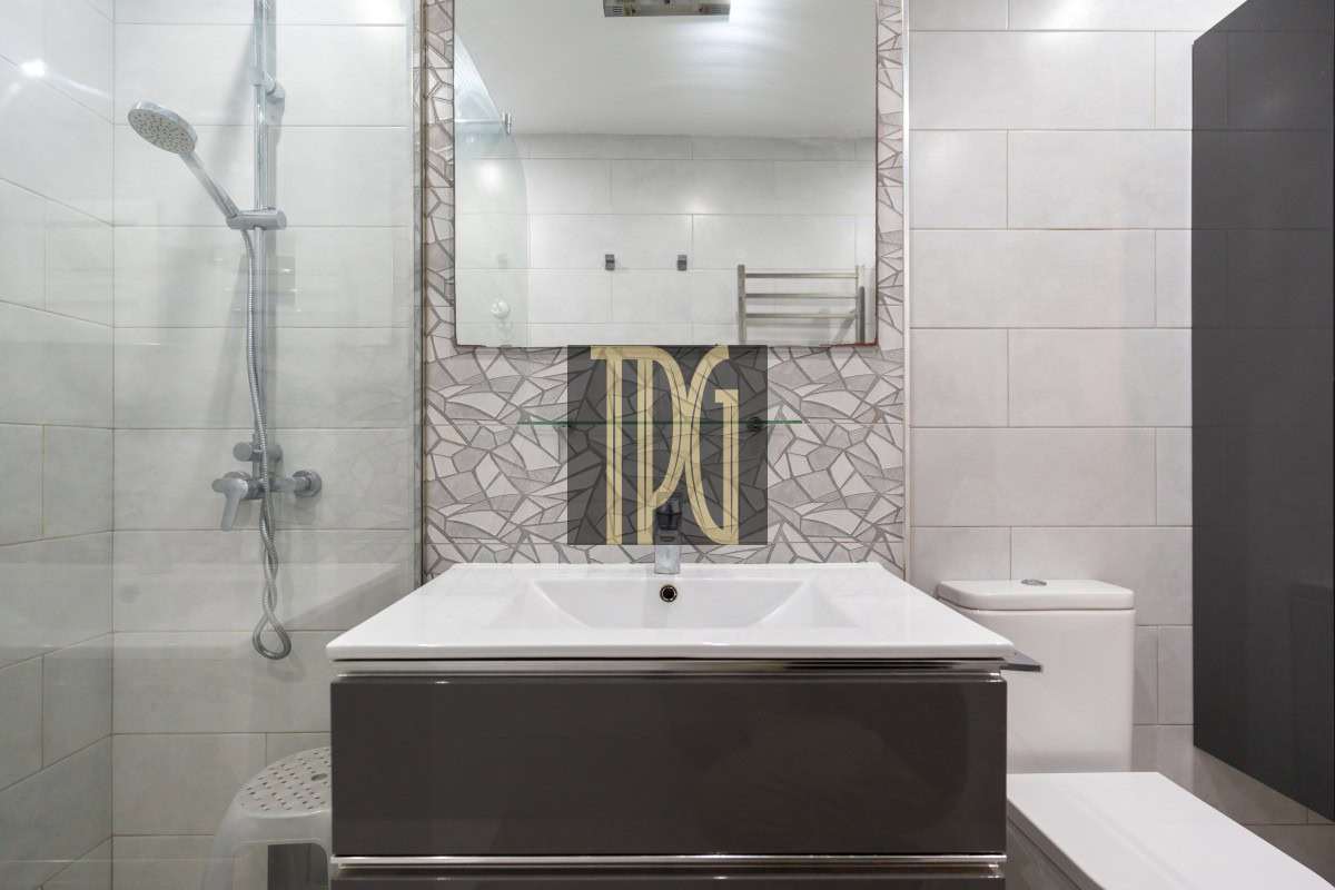 Modern bathroom with a glass shower on the left, a white sink on a dark vanity, and a geometric tiled backsplash behind the basin.