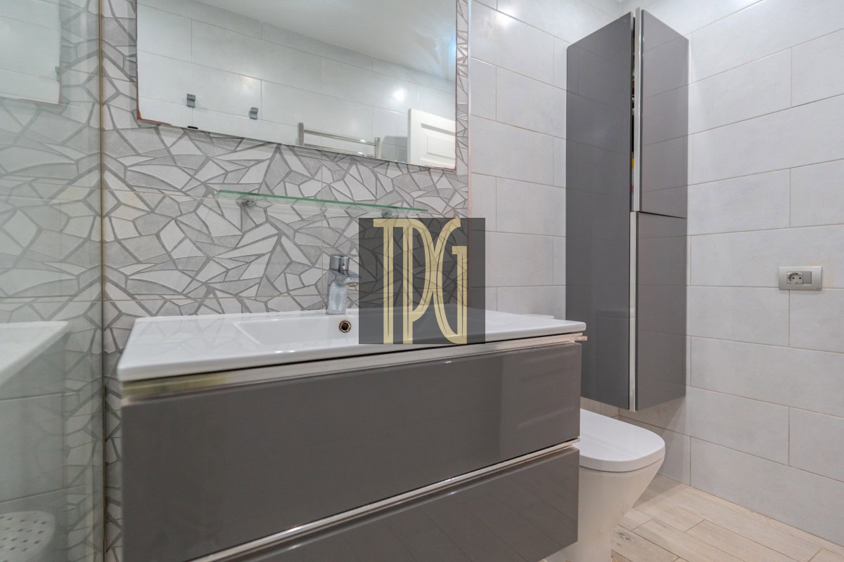 Modern bathroom with a glossy gray vanity, white sink, large mirror, geometric tiled backsplash, wall shelf, and toilet nearby.