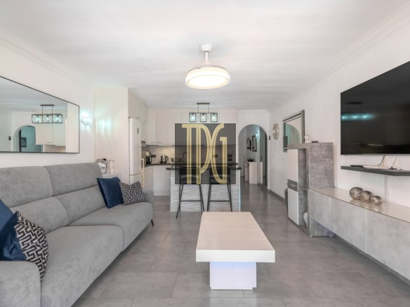Open-concept living room and kitchen with a gray sofa, blue cushions, and a large wall-mounted TV across from a tiled coffee table.