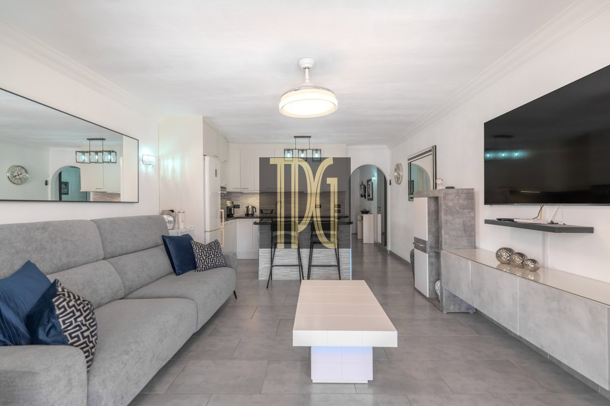 Open-concept living room and kitchen with a gray sofa, blue cushions, and a large wall-mounted TV across from a tiled coffee table.