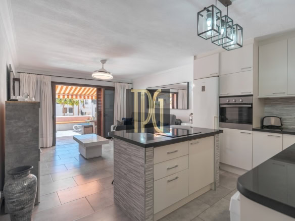 Open-plan kitchen with a central island, white cabinets, and a view into the living area and outdoor patio with an orange-striped awning.
