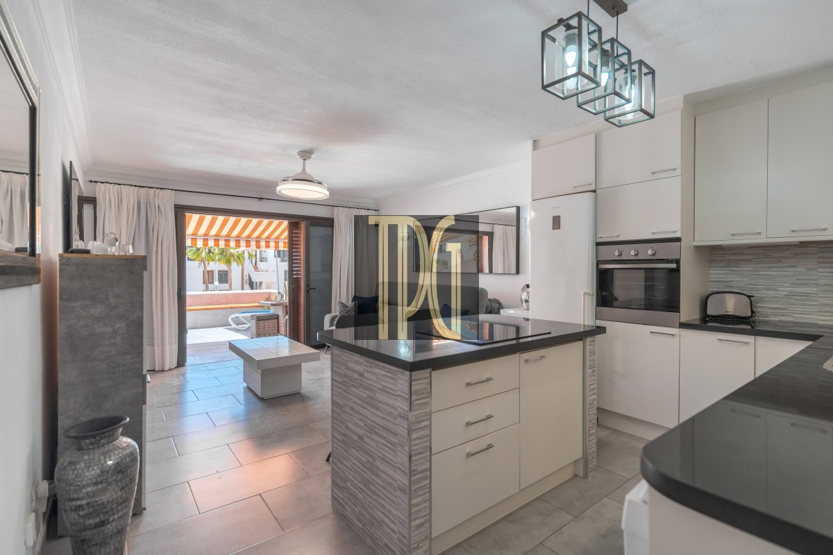 Open-plan kitchen with a central island, white cabinets, and a view into the living area and outdoor patio with an orange-striped awning.