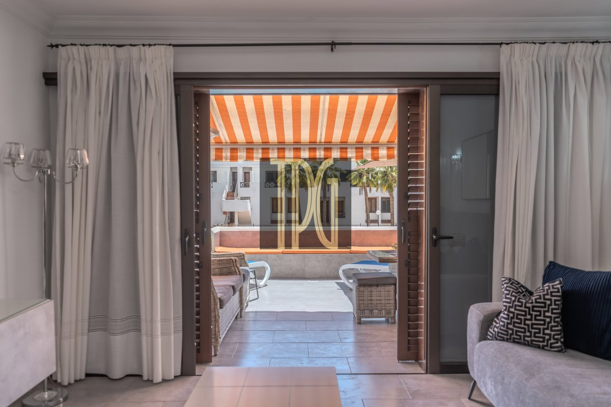 Living room opens to a sunlit terrace with an orange-striped awning, wicker chairs, and outdoor seating.