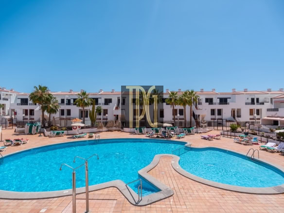 Outdoor hotel pool area with curved blue water pool, tan tile deck, and lounge chairs against white apartment buildings and palm trees.