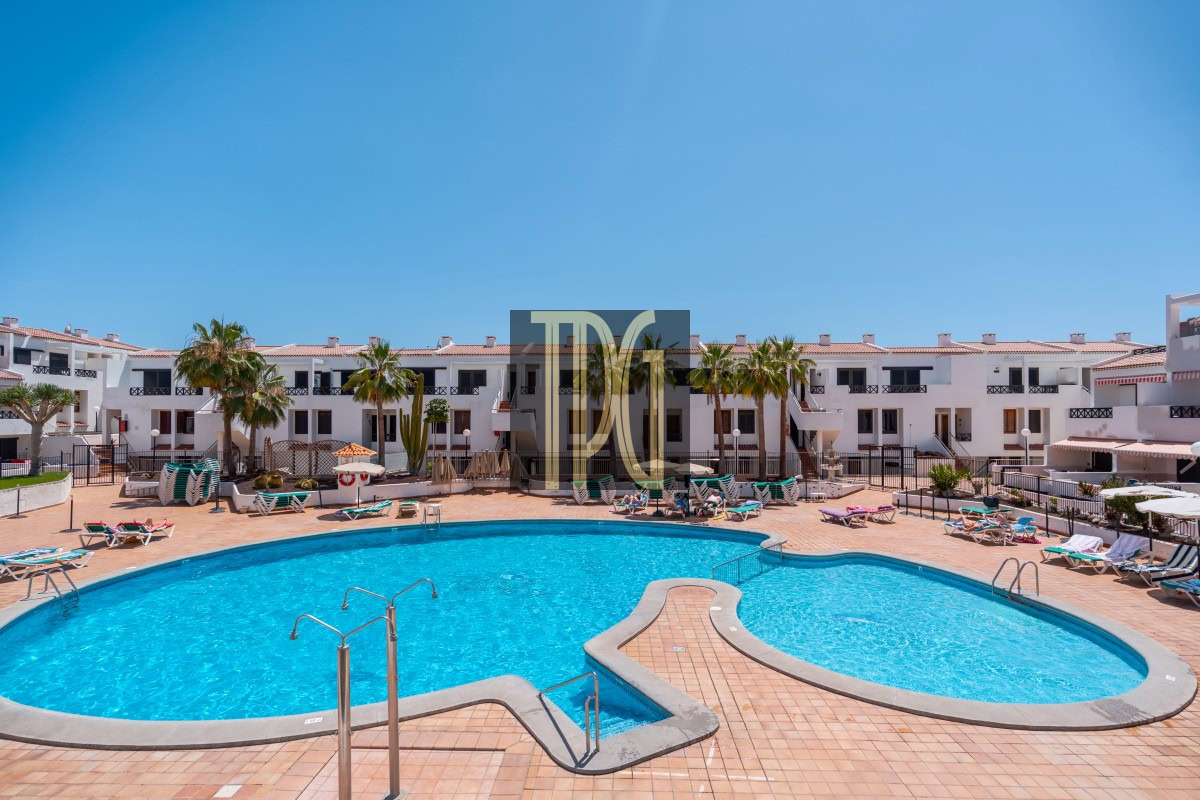 Outdoor hotel pool area with curved blue water pool, tan tile deck, and lounge chairs against white apartment buildings and palm trees.