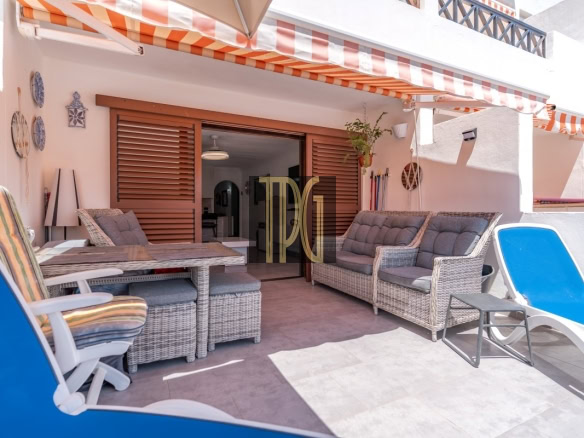 Outdoor patio with wicker seating, a glass tabletop, blue lounge chairs, and orange-striped awnings over a sliding wooden door to the house.