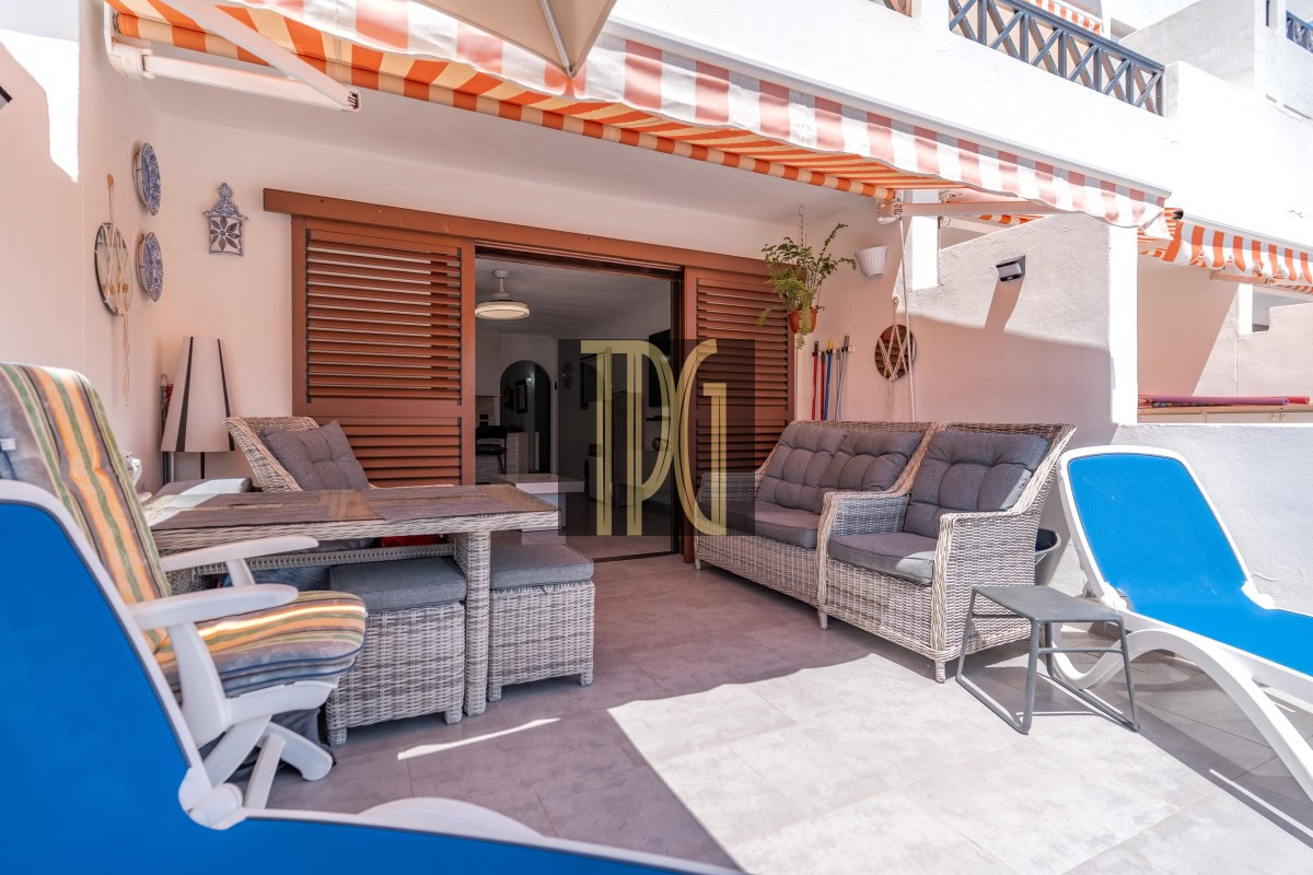 Outdoor patio with wicker seating, a glass tabletop, blue lounge chairs, and orange-striped awnings over a sliding wooden door to the house.