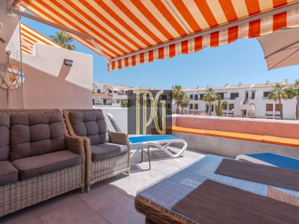 Sunny rooftop terrace with an orange-striped awning, wicker seating, and sun loungers under a blue sky.