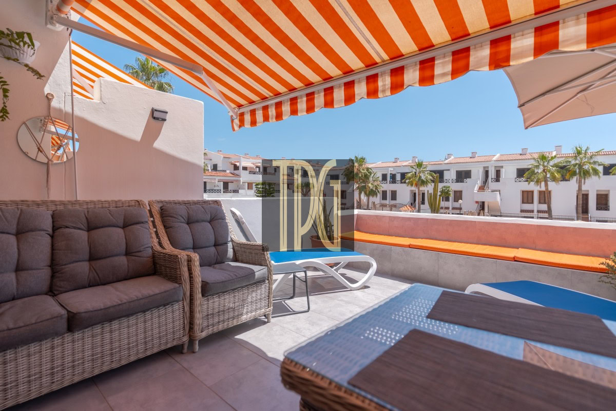 Sunny rooftop terrace with an orange-striped awning, wicker seating, and sun loungers under a blue sky.