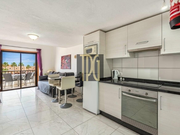 Open-plan kitchen and living area with white cabinets, black countertops, bar stools, and a balcony with seating and palm trees outside.