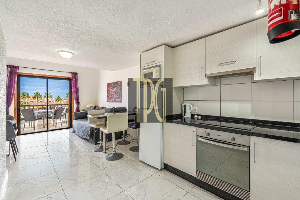 Open-plan kitchen and living area with white cabinets, black countertops, bar stools, and a balcony with seating and palm trees outside.