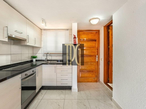 Modern L-shaped kitchen with white cabinets, black granite countertops, and a built-in oven beside a sink under a window with blinds, near a wooden entry door.