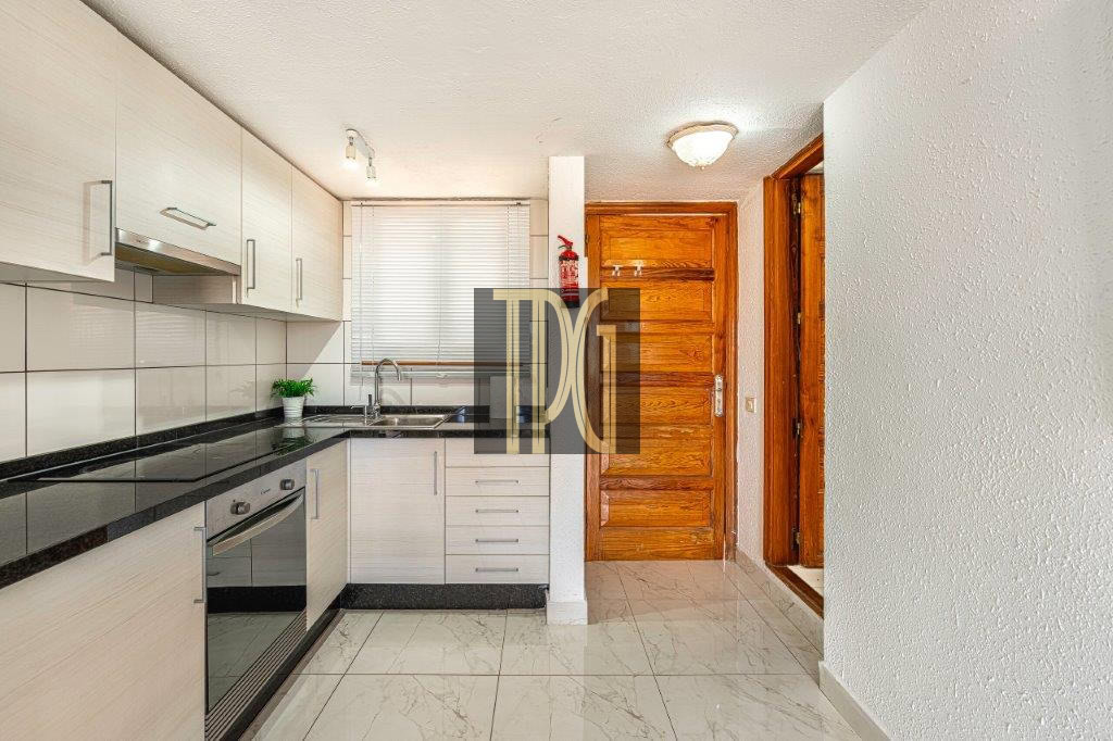 Modern L-shaped kitchen with white cabinets, black granite countertops, and a built-in oven beside a sink under a window with blinds, near a wooden entry door.
