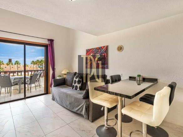 Living area with a gray sofa, bar-height dining table and cream stools, and a sliding glass door to a balcony with outdoor furniture and palm trees outside.